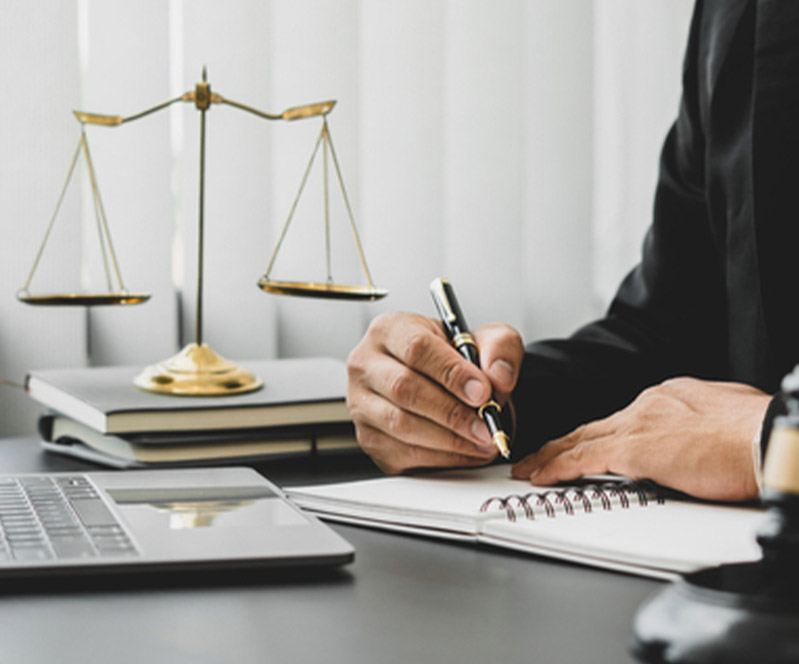 Legal professional taking notes at a desk with a scale of justice and a laptop.