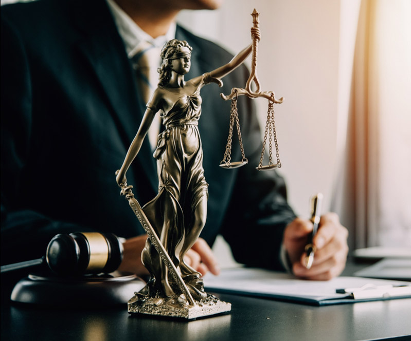Lady Justice statue on a desk with a lawyer and gavel in the blurred background.